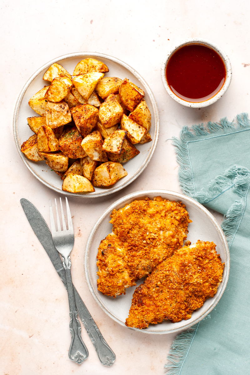 Parmesan crusted chicken breasts on a plate and air-fried potatoes on another. 
