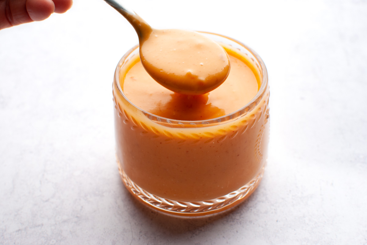 Bang bang sauce in a clear glass jar with a spoon full of sauce positioned over it. 